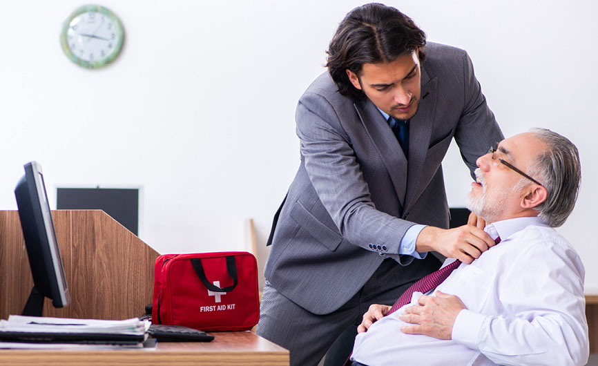 Office worker providing first aid to an older colleague, loosening his tie while a red first aid kit is placed on the desk.