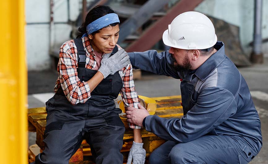 Worker in a safety helmet assisting a colleague holding her shoulder, both in industrial attire, with tools and machinery in the background.