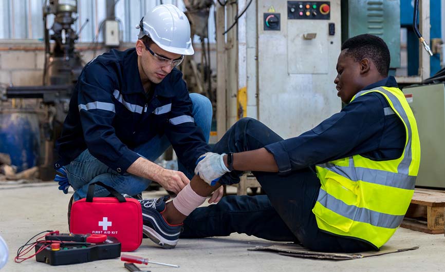 Industrial worker in safety gear applying a bandage to a coworker’s injured leg, with a red first aid kit visible in the workshop.