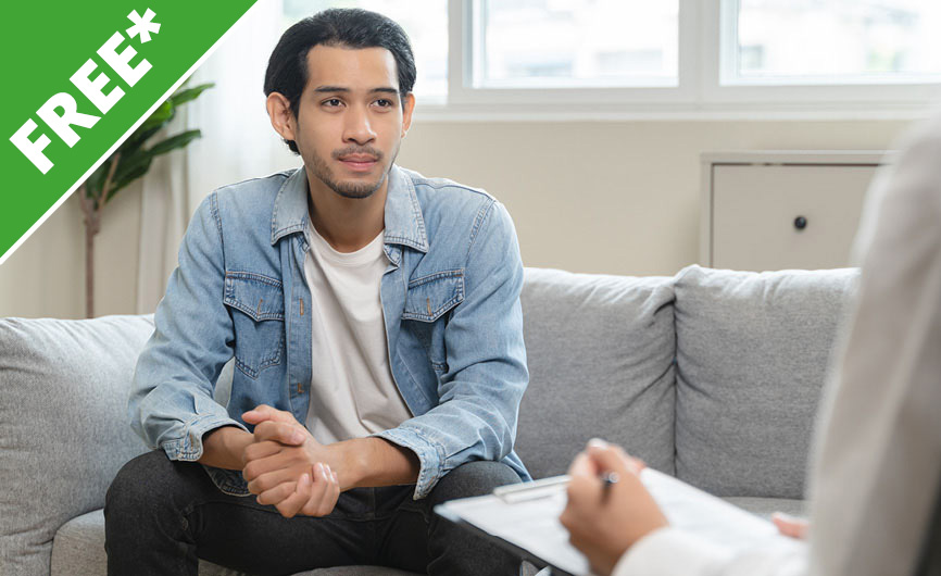 A man sitting on a couch during a mental health consultation, attentively listening to a counselor whose notepad is partially visible.