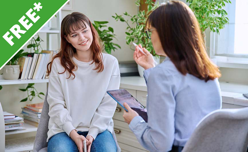 A young woman smiling and engaging with a mental health counselor holding a tablet, in a cozy and well-lit office with plants and shelves in the background.