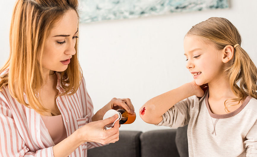 A woman applying ointment to a young girl's scraped elbow, while the girl smiles and sits calmly on a couch.